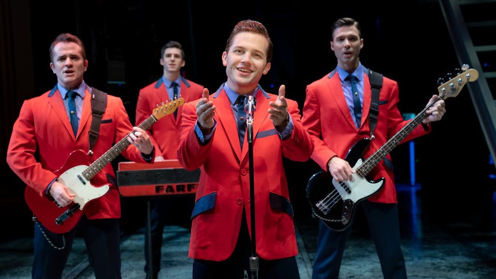 The four principal members of Jersey Boys standing and singing in red jackets. Frankie Valli is in the foreground with his arms outstretched looking at the camera