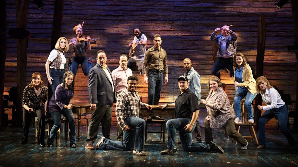 The cast of Come from Away standing and kneeling around a wooden table and looking at the camera
