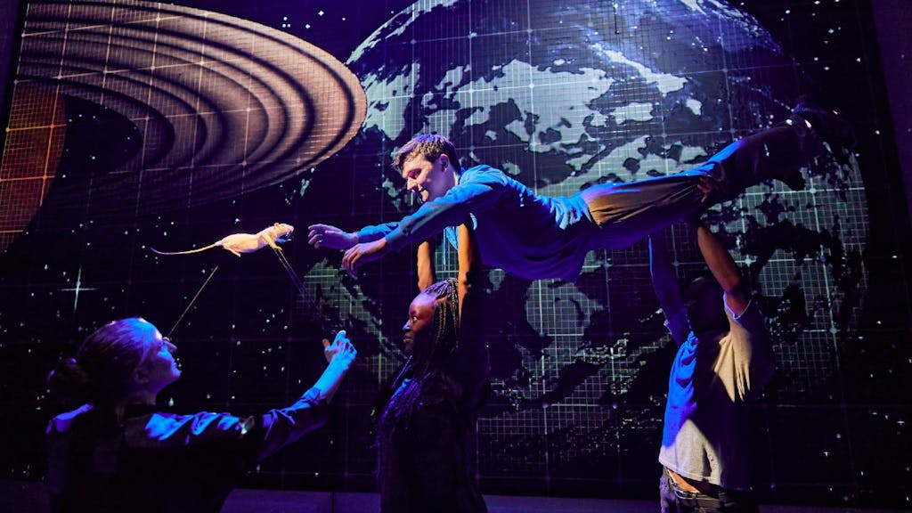 From The Curious Incident of the Dog in the Night-Time, the main character is held up by another actor and has his arm outstretched to a small rat. Planets are visible in the background