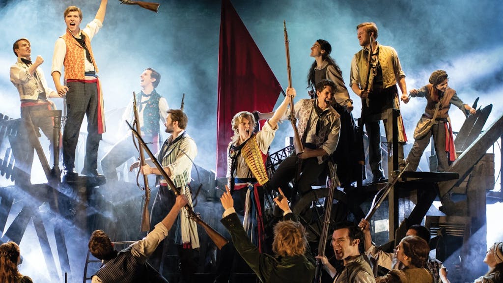 the ensemble of les miserables standing on barricades in front of a large red flag